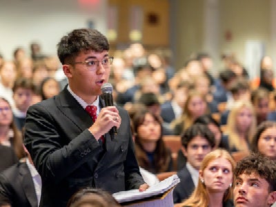 A student wearing a suit stands with a microphone in a full auditorium.