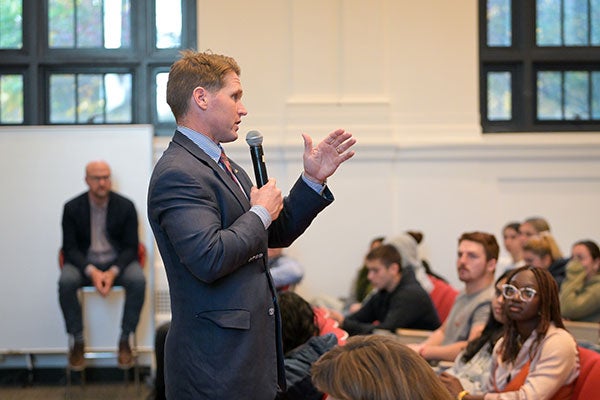 A man standing in front of a lecture hall holding a microphone and speaking to students.