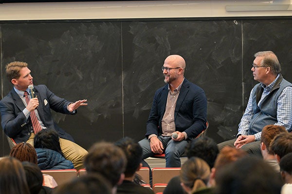 3 men sitting on chairs in front of a class, one is speaking and gesturing and the other two are looking at him.