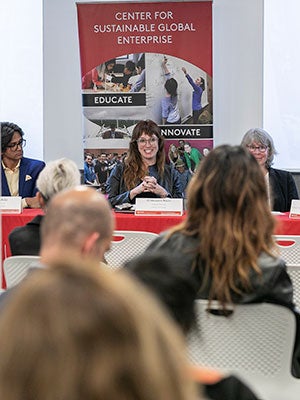 A woman speaking, seated next to other panelists at a table at the front of a room with the backs of the heads of the audience in the foreground.
