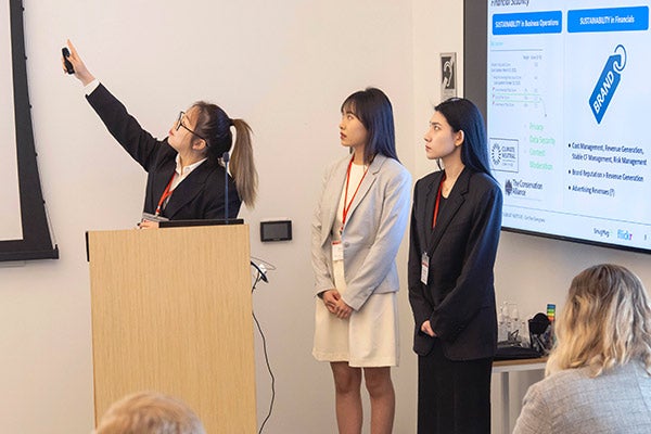 one woman standing behind a podium points up at a screen while the other two women look on. The backs of two audience members&rsquo; heads are in the foreground.