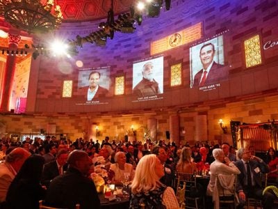 Photo of an elegant restaurant with a high ceiling and a huge chandelier hanging high above all. The room is filled with people sitting at tables and images of the evening’s honorees are projected onto the stone walls above the room’s columns.