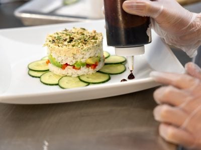 A gloved hand squeezes dots of sauce around a dish of rice and vegetables.