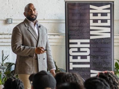 A man standing in front of a room full of people next to a Tech Week banner and speaking.