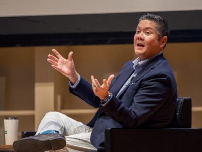 A man sits on stage talking with his hands outstretched.