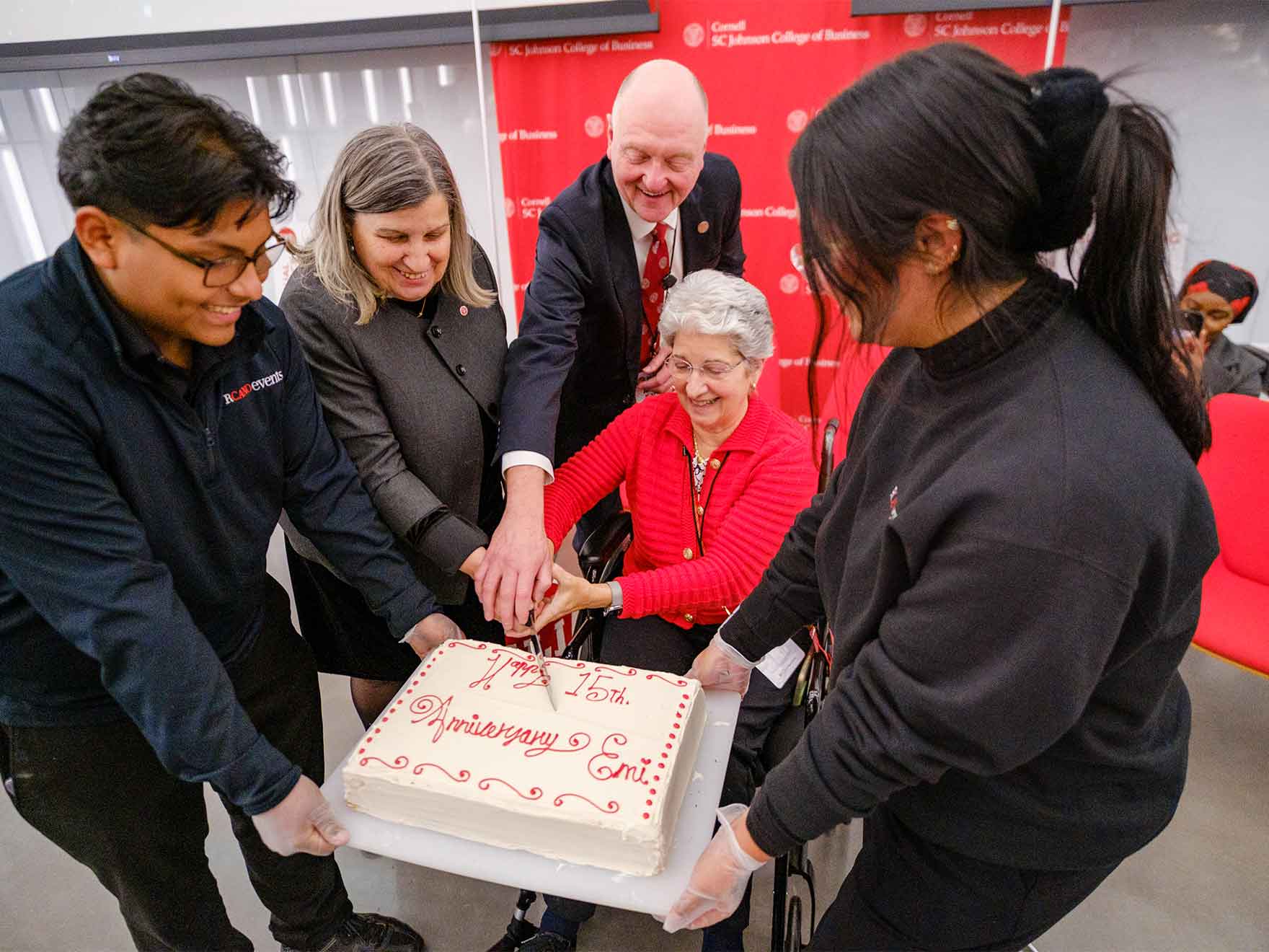 Two people hold a big cake while three others hold the knife, poised to cut.