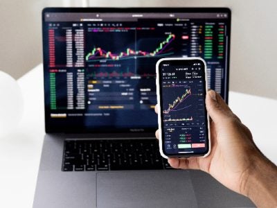 Image of a human hand holding a cell phone in front of a laptop with both displaying stock market activities