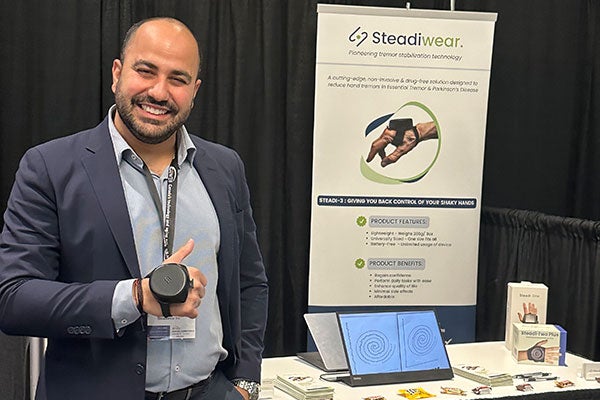 A man wearing a round device attached to the back of his hand and standing next to a table and poster displaying information about Steadiwear.