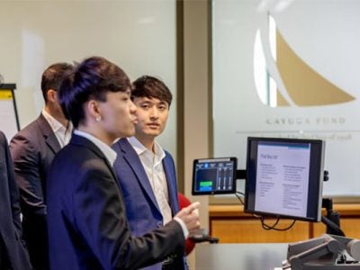 A male student standing in front of a podium and screen addresses an audience while another male student standing next to him looks on. Both are dressed in business suits.