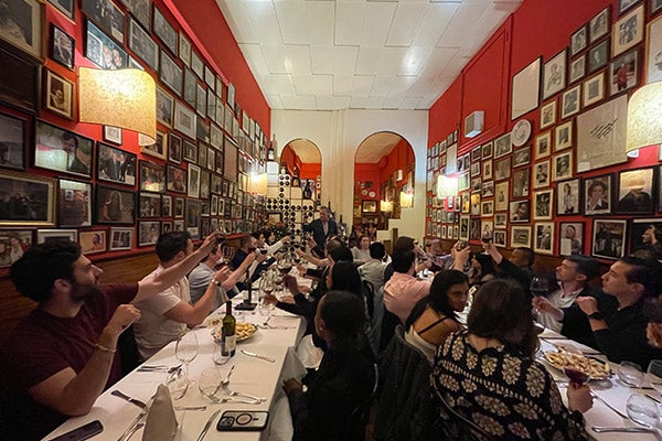 A large group group of students seated at long tables raise their glasses in a toast.