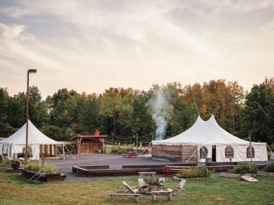 Two large tents one either side of an open area with a fire pit and wooden chairs in the foreground.
