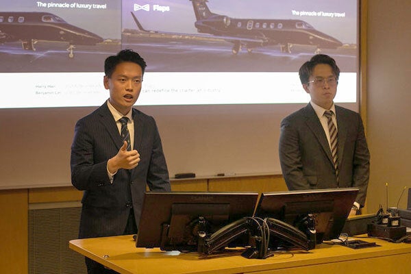 Two men standing in front of dual screens depicting airplanes. One is speaking and gesturing.