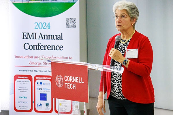 A woman standing at a podium and speaking into a microphone next to a sign with the text: 2024 EMI Annual Conference.