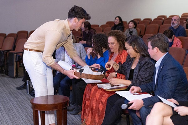 A young man holding a tray leans down to present it to several people sitting in the front row of an auditorium.