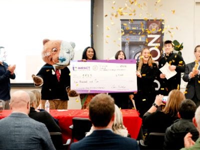 Students stand with a bear mascot and an oversized check in front of an audience as gold confetti falls.