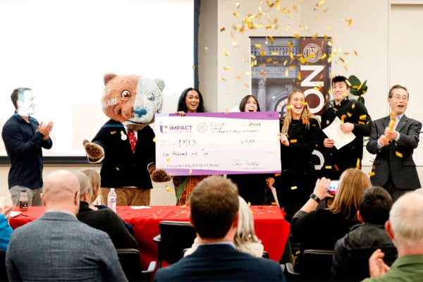 Students stand with a bear mascot and an oversized check in front of an audience as gold confetti falls.