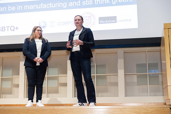 Two women stand on a stage as the woman on the right gestures and addresses the audience.