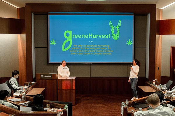 two women speaking at the front of a tiered room with people sitting at rows of built-in wooden tables with a screen that says &ldquo;green harvest&rdquo; in the background.
