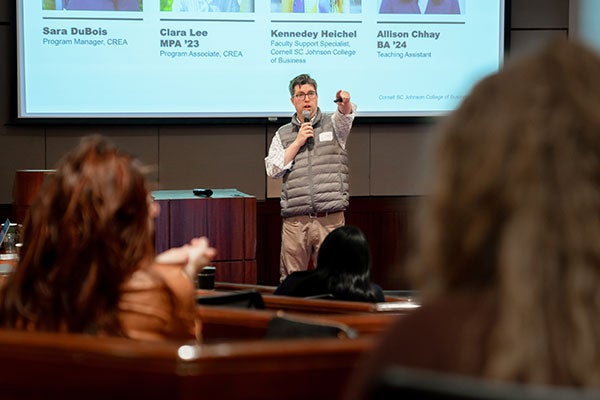 a man holding a microphone stands at the front of a room, with a screen in the background, speaking and gesturing.