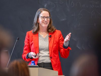 A woman standing at a podium in front of a classroom of students, speaking, smiling, and gesturing with one hand.
