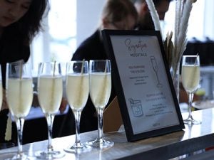 A row of light yellow cocktails on a counter top next to a menu card listing its ingredients.