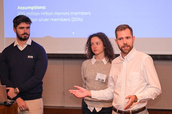 Three students dressed in business casual standing in front of a screen. Two are listening, one is speaking and gesturing.