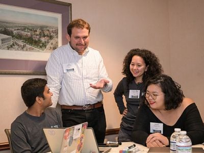 Four students dressed in business casual, two seated and two standing, around a table and talking while looking at a laptop screen.