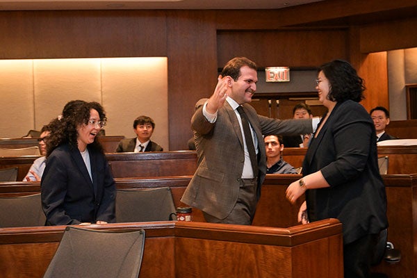 Students dressed in business attire standng and smiling; one is reaching out arms to hug another and a third team member is right behind. They are in an auditorium.