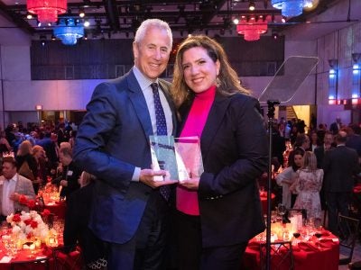 Award-winners Mia Kyricos, MMH ‘03, and Danny Meyer stand on stage at the 14th annual Icons and Innovators Award ceremony, with the crowd and tables behind them.