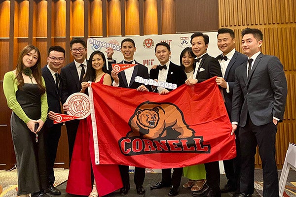 A group of 3 women and 7 men dressed in formal attire smiling and holding a Cornell banner displaying the Cornell bear.