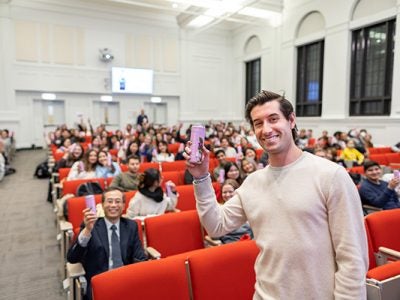 Steven Izen standing in front of a 2/3 full lecture hall, holding up a can and smiling. Many audience members are holding up similar looking cans.