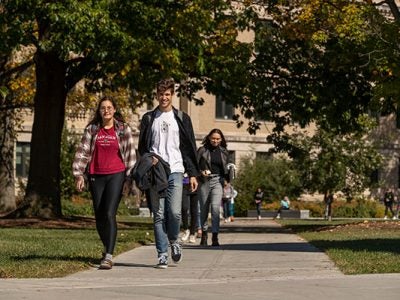 Students walking on sidewalk paths under a canopy of trees with a college building in the background.
