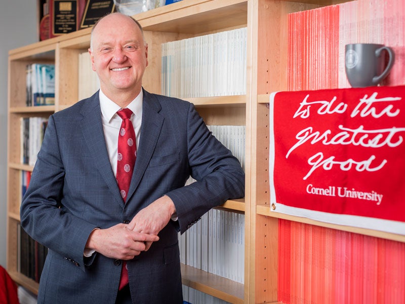 Portrait of Dean Andrew Karolyi, standing in office next to a "To do the greatest good" banner