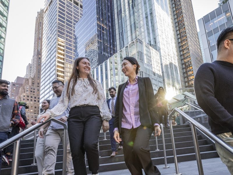Two young women in business casual attire, along with other students in Cornell's Baker Program in Real Estate, stroll down an outdoors granite staircase with stainless steel railing, against a sun-reflective backdrop of several large contemporary high-rise buildings in New York City