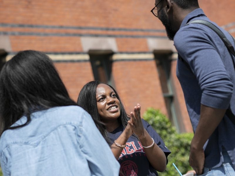 Two young women and a young man, all enrolled in the Master of Professional Studies in Management program at Cornell's Johnson School, talk outdoors in the sun against a backdrop of a brick building