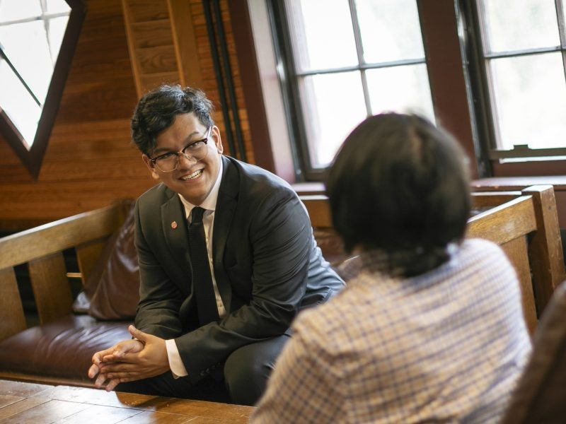 Two men, students in the master's program in accounting at Cornell's Johnson School, meet and discuss coursework in business formal attire in a well-lit wood-walled cafe