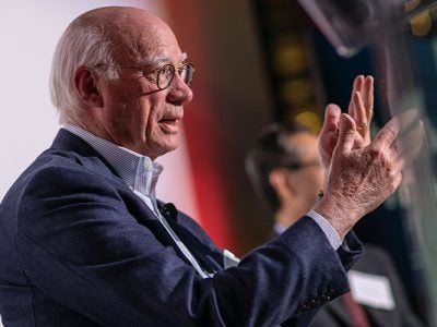 A close-up photo of a man’s head and hands as he gestures with his hands and speaks.