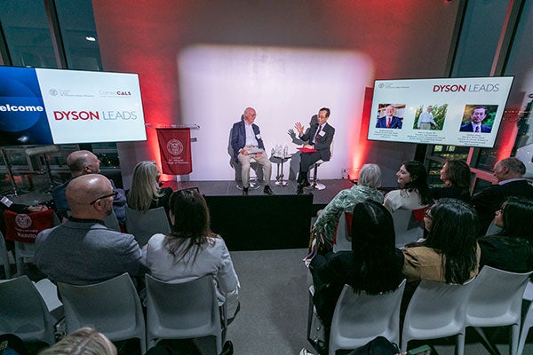 In a photo taken from the back of the room, two men seated on stage and speaking to a room full of people.