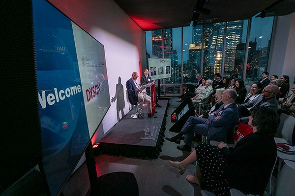 A photo taken from the side of a stage with two men seated and a room full of people smiling and laughing with floor to ceiling windows revealing a night time cityscape in the background.