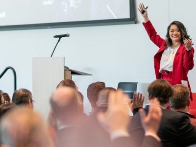Gage Javier stands on stage in a red business suit with her arm raised, and audience members in the foreground also raise their hands.