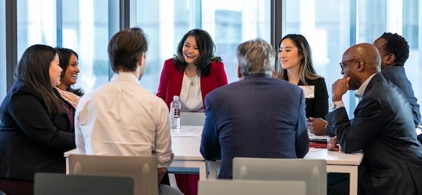 A group of alumni chat at a table in a conference room with large windows.