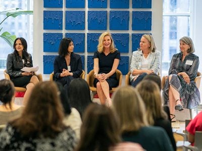 5 women sitting on a low, platform stage with the heads of the audience in the foreground.