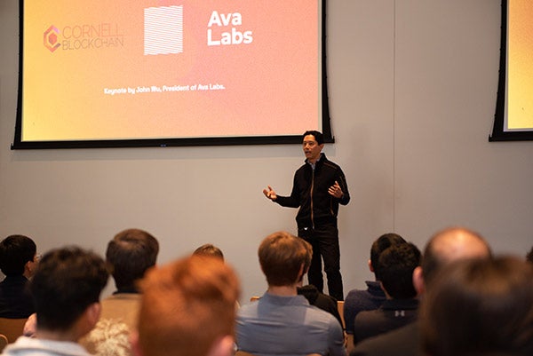 John Wu standing and speaking on a stage at the front of a lecture hall / large room filled with students.