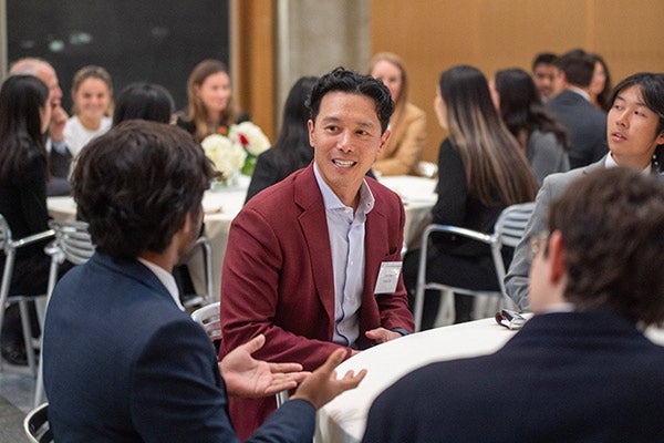 John Wu sitting at a table talking to students with other tables of students seated at tables in the background.