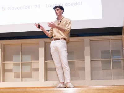 A young man standing on a stage, gesturing with both hands, and addressing the audience.