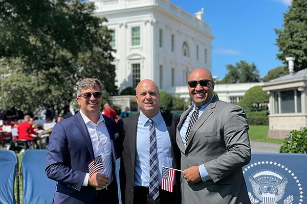 Three men standing side by side, holding flags, with the White House in the background.
