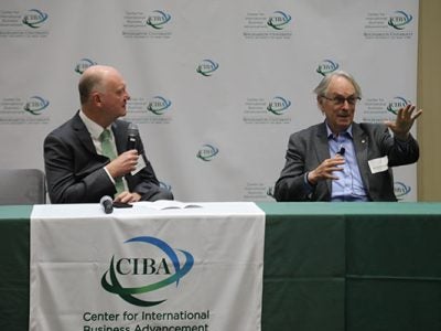 Andrew Karolyi and Stanley Whittingham seated at a table draped with a CIBA banner and speaking.