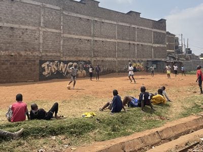 children playing soccer in the dirt in front of a large brick wall.