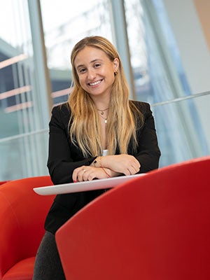 envionmental photo of Megan Shkolyar seated in a bright red char with anohter red chair in the foreground and windows in the background.,
