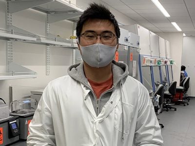 Masahiro standing in a white lab coat in front of metal shelving.